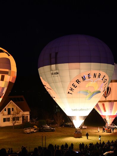 Colorful hot air balloons light up the darkness and attract many spectators. In the background, a house and a crowd of people can be seen.