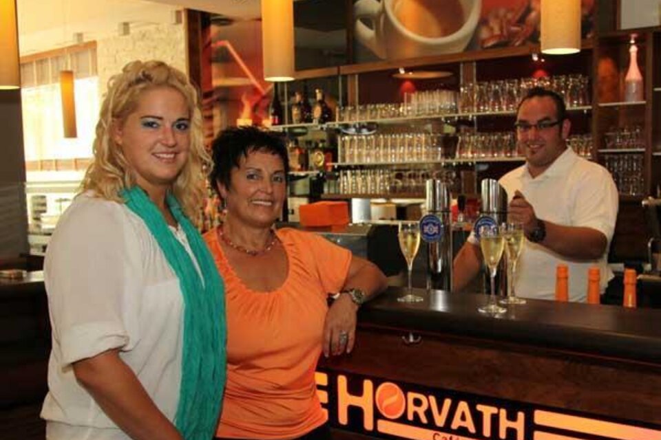 Eine Bar mit einer modernen Einrichtung. Zwei Frauen stehen im Vordergrund, während ein Bartender im Hintergrund arbeitet. | © Cafe Horvath