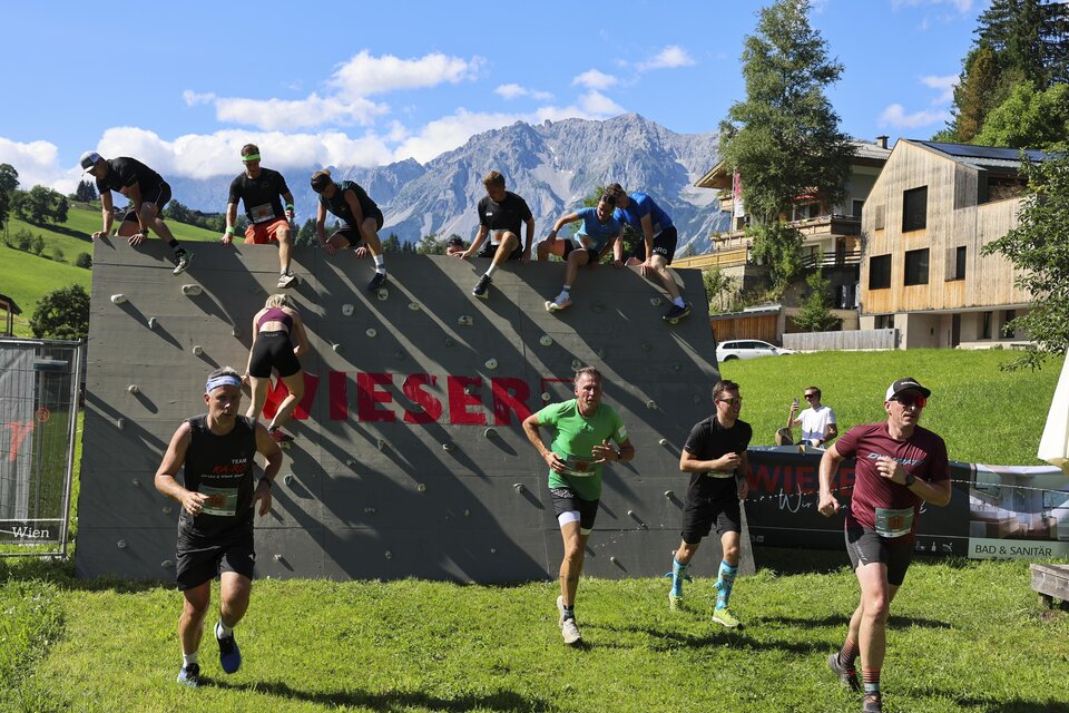 Eine Gruppe von Sportlern nimmt an einem Hindernislauf teil und überwindet eine Kletterwand. Im Hintergrund sind Berge und Gebäude in einer malerischen Landschaft zu sehen. | © Wolfgang Grebien