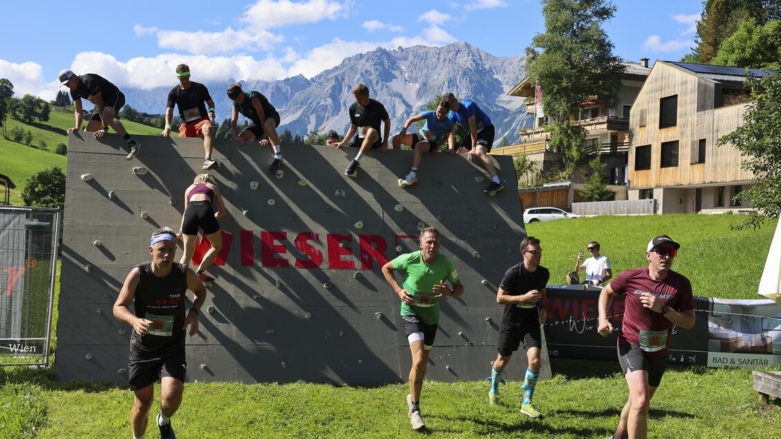 Eine Gruppe von Sportlern nimmt an einem Hindernislauf teil und überwindet eine Kletterwand. Im Hintergrund sind Berge und Gebäude in einer malerischen Landschaft zu sehen. | © Wolfgang Grebien