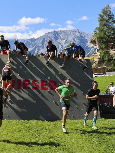 Eine Gruppe von Sportlern nimmt an einem Hindernislauf teil und überwindet eine Kletterwand. Im Hintergrund sind Berge und Gebäude in einer malerischen Landschaft zu sehen. | © Wolfgang Grebien