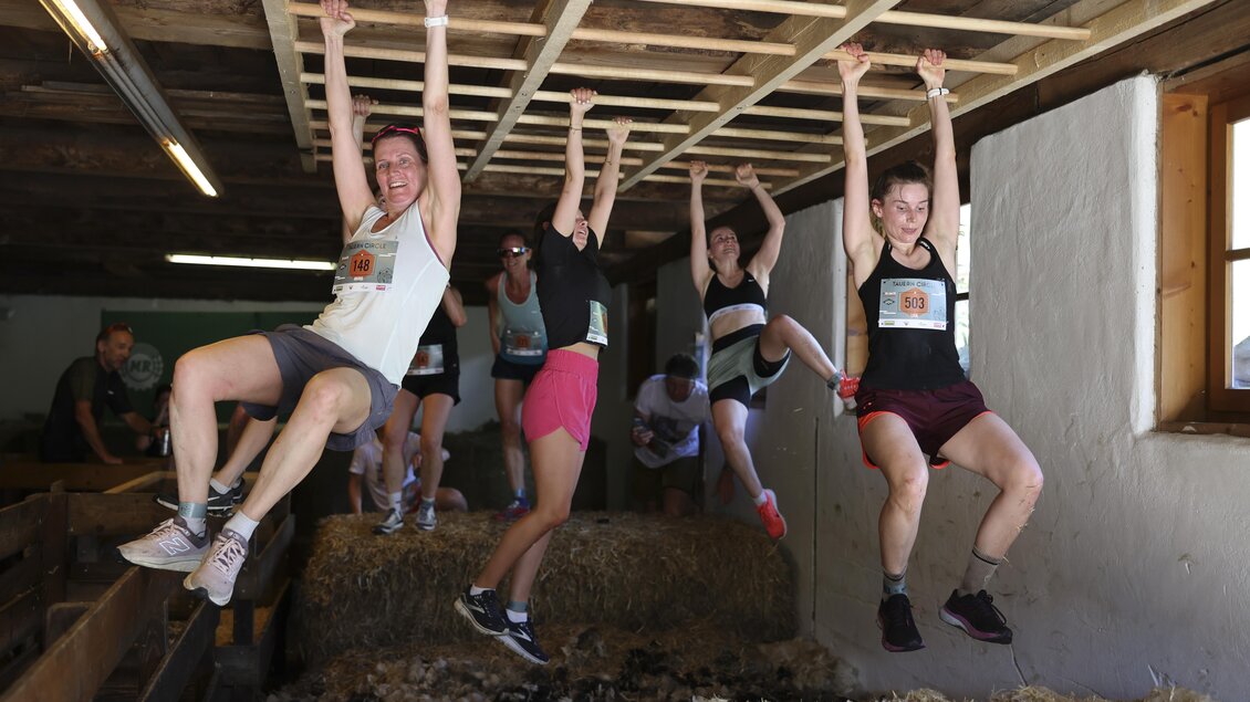 Eine Gruppe von Menschen hängt an einem Balken in einem Stall. Sie sind aktiv und scheinen an einem Sport- oder Fitnessereignis teilzunehmen. | © Wolfgang Grebien