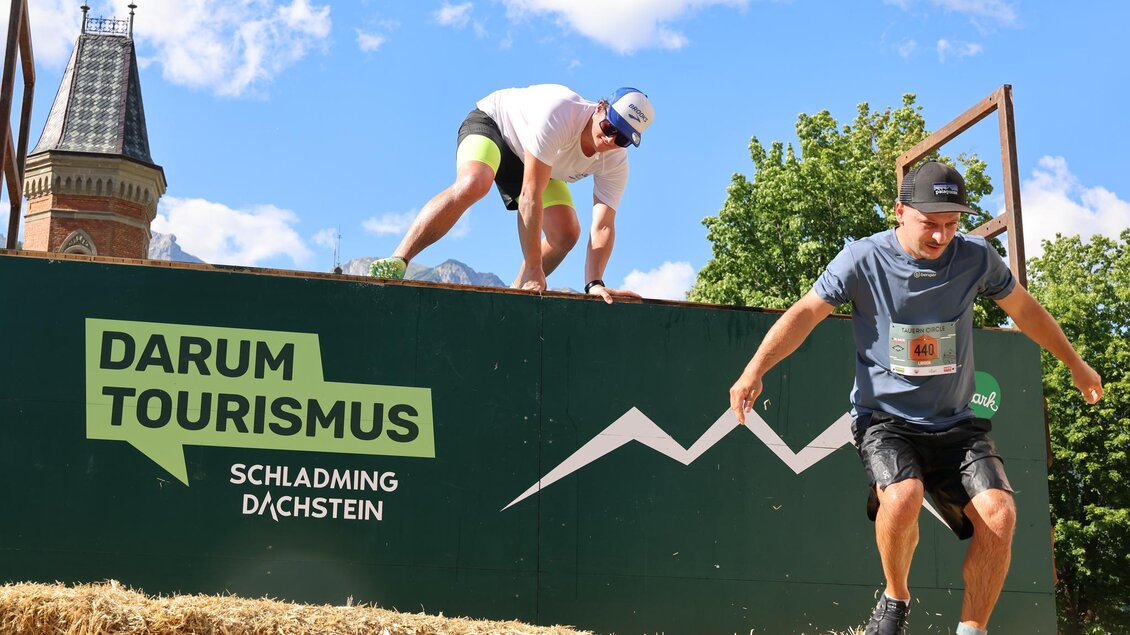 Zwei Männer springen von einer Plattform und nehmen an einem Sportevent teil. Im Hintergrund ist ein Werbeschild für den Tourismus in Schladming-Dachstein zu sehen. | © Michael Simonlehner