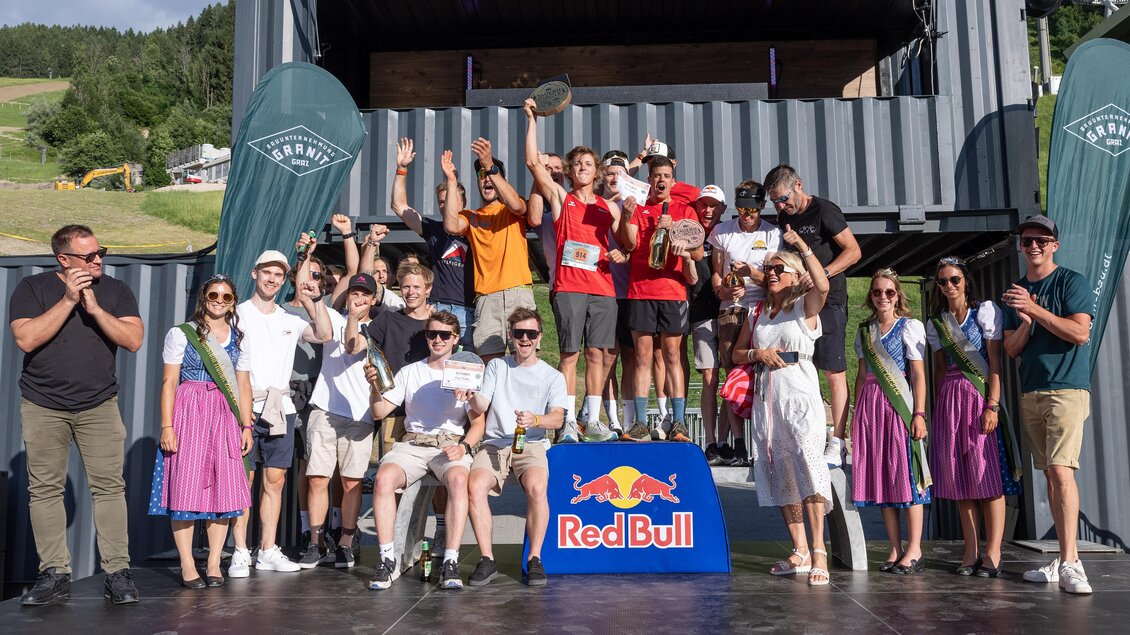Eine Gruppe von fröhlichen Teilnehmern steht auf einer Bühne und hält Pokale in den Händen. Im Hintergrund sind Berge und Banner der Marke Red Bull zu sehen. | © Harald Steiner