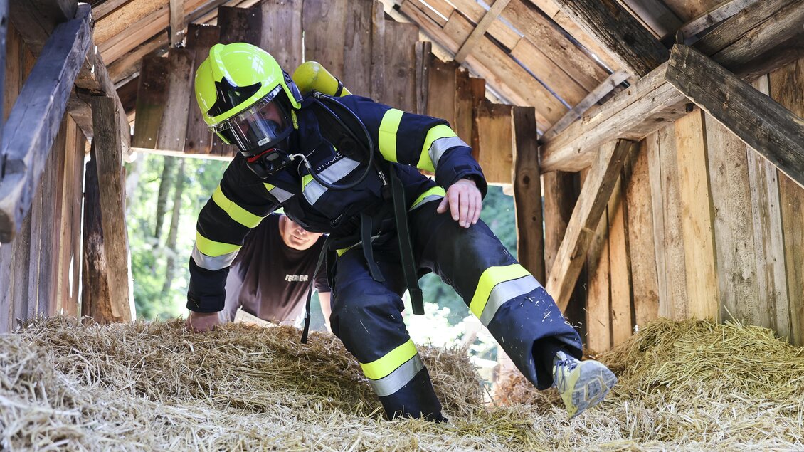 Ein Feuerwehrmann klettert in eine Scheune voller Stroh. Im Hintergrund ist eine weitere Person, die ihn beobachtet. | © Wolfgang Grebien
