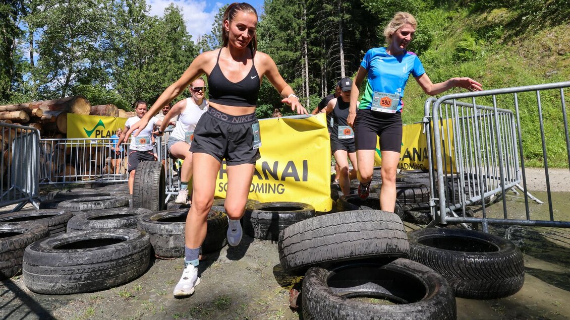 Eine Gruppe von Frauen überwinden Hindernisse mit Autoreifen während eines Outdoor-Events. Im Hintergrund sind Bäume und eine gelbe Sponsorentafel zu sehen. | © Michael Simonlehner