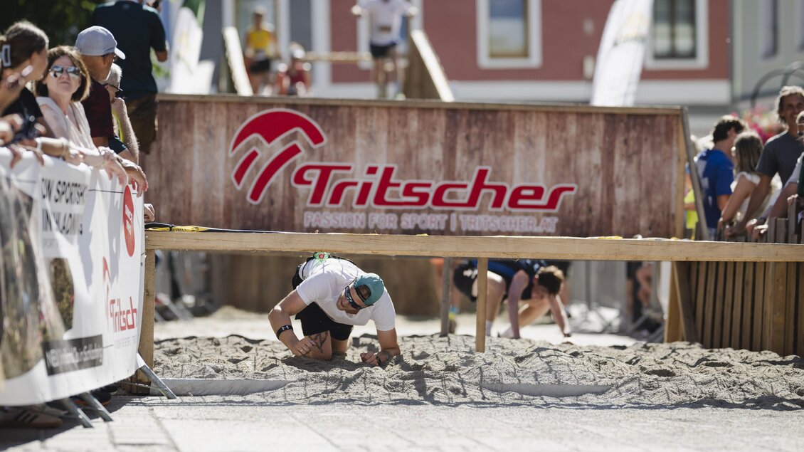 Ein Wettkampf, bei dem Teilnehmer unter einem Hindernis durchkriechen. Zuschauer beobachten die Veranstaltung gespannt. | © Simone Ranninger