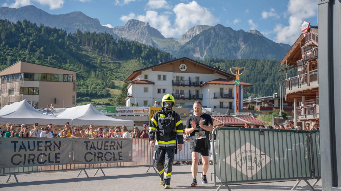 Ein Feuerwehrmann begleitet einen Läufer durch das Ziel bei einem Wettkampf. Im Hintergrund sind die Berge und eine große Menschenmenge zu sehen. | © Simone Ranninger