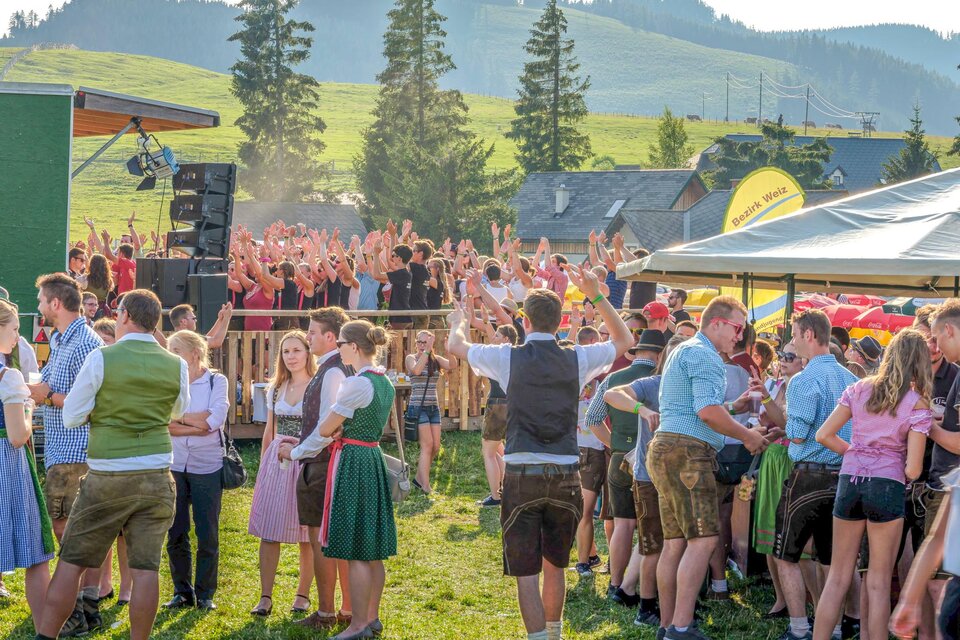 Eine fröhliche Menschenmenge auf einer Wiese, die in traditioneller Kleidung tanzt und feiert. Im Hintergrund sind Bäume und eine Bühne zu sehen. | © Fotoglick