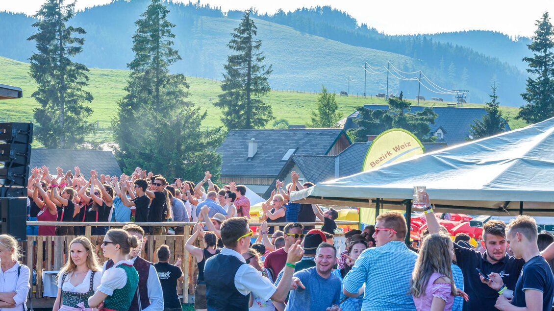 Eine fröhliche Festveranstaltung im Freien mit vielen Menschen, die tanzen und feiern. Im Hintergrund sind grüne Hügel und Bäume sichtbar. | © Fotoglick