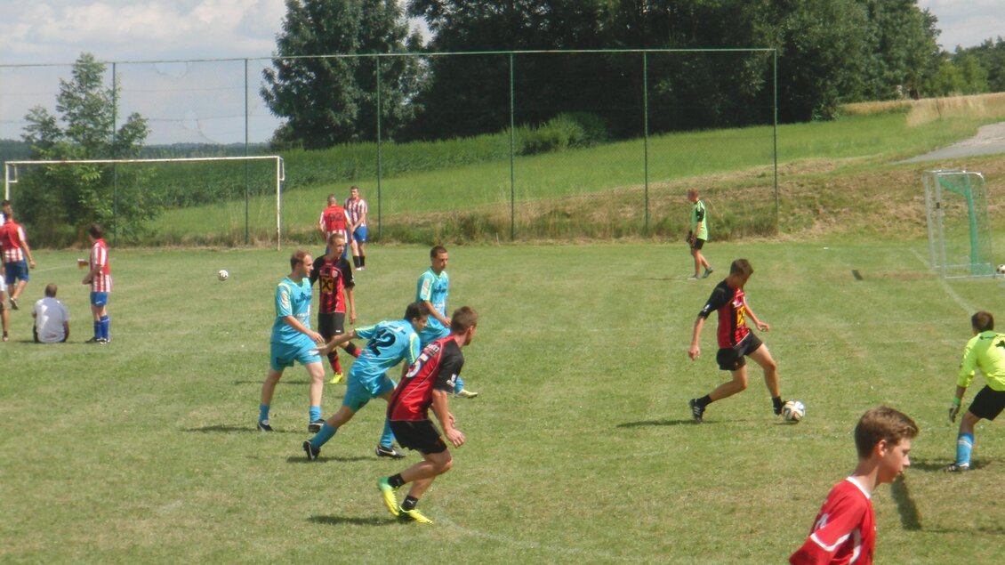 Eine Fußballspielszene mit mehreren Spielern auf dem Feld. Im Hintergrund sind Bäume und ein Fußballtor zu sehen. | © Roland Hofer