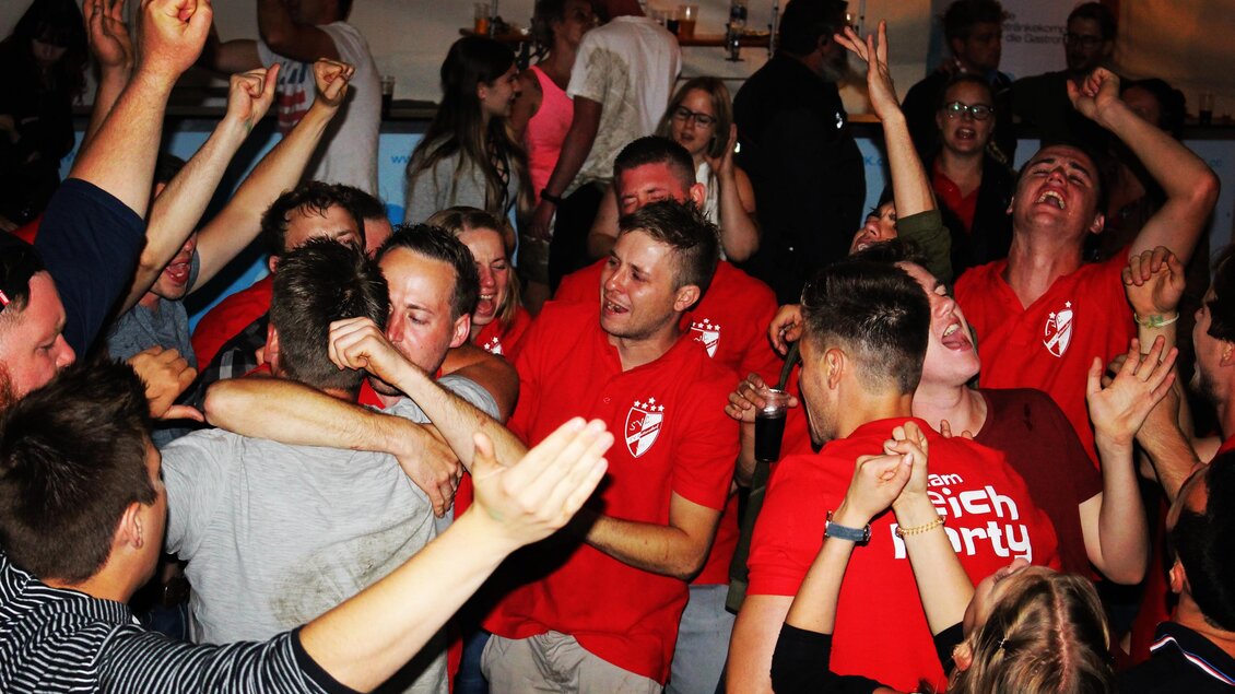 Eine lebhafte Feier mit jubelnden Menschen in roten T-Shirts. Die Leute tanzen und feiern zusammen in einer fröhlichen Atmosphäre. | © Roland Hofer