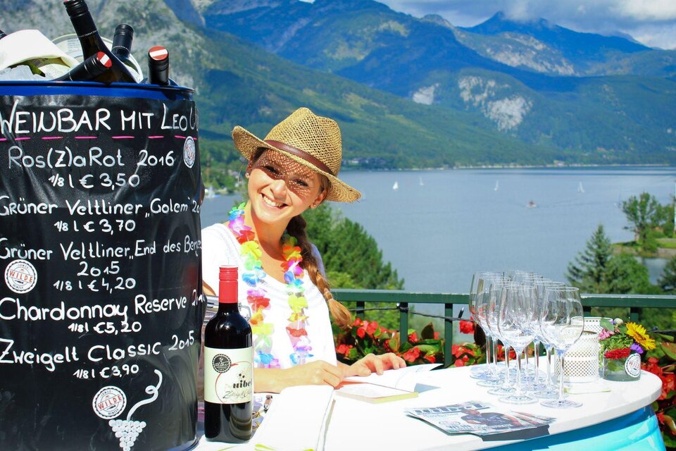 Eine fröhliche Frau sitzt an einer Weinstube mit Blick auf einen See und Berge. Auf dem Tisch stehen Weingläser und verschiedene Weinsorten sind auf einer Tafel aufgelistet. | © Mondi Resort