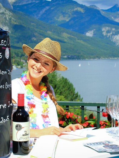 A cheerful woman sits at a wine tavern with a view of a lake and mountains. On the table, there are wine glasses and various types of wine are listed on a board. | © Mondi Resort