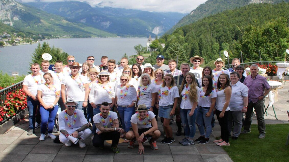 Eine Gruppe von Menschen steht lächelnd zusammen am Ufer eines Sees. Im Hintergrund sind Berge und eine malerische Landschaft zu sehen. | © Mondi Grundlse