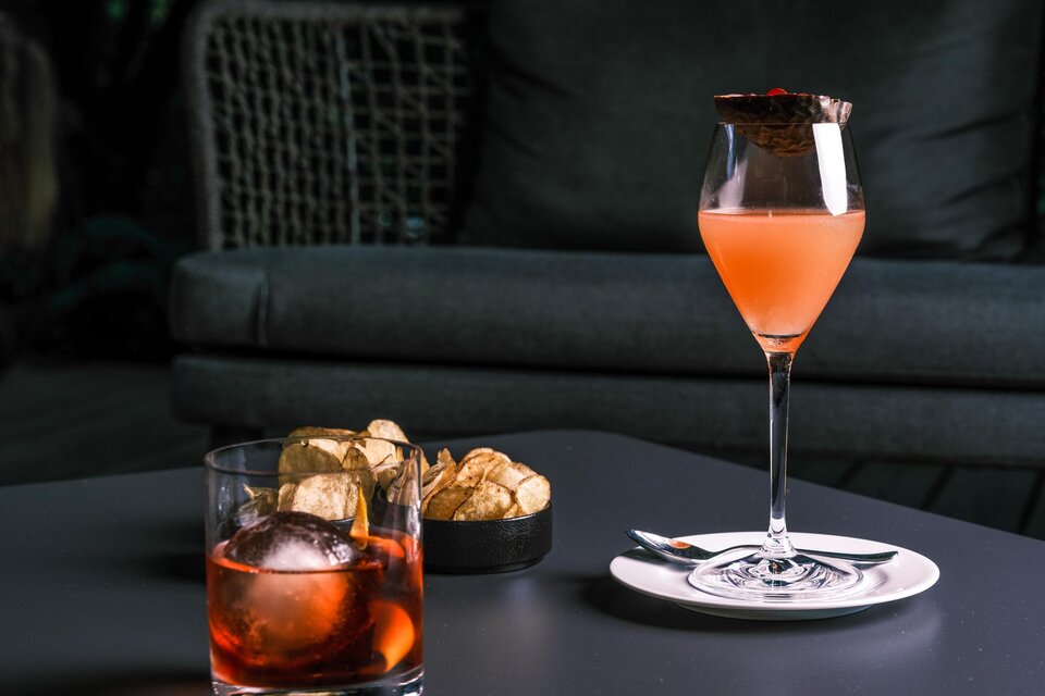 Ein eleganter Cocktail in einem Glas mit einem fruchtigen Farbton steht auf einem Tisch. Daneben befinden sich ein Glas mit einem Getränk und eine kleine Schale mit Snacks. | © Falkensteiner Hotel Schladming / Fotograf Dragan Dokmanovic