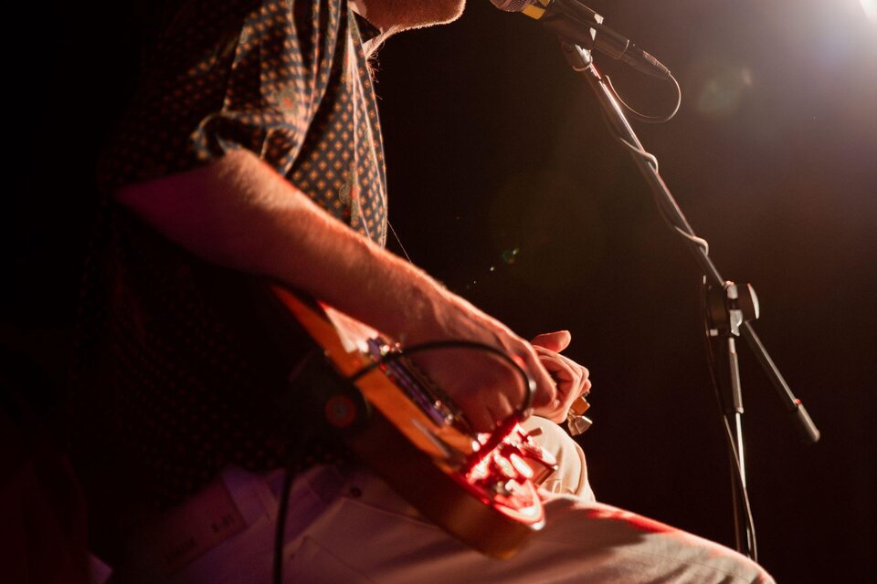 Ein Musiker sitzt auf einem Hocker und spielt ein Instrument. Er singt ins Mikrofon und ist auf einer Bühne beleuchtet. | © TVB Südsteiermark–Achromatic Photography