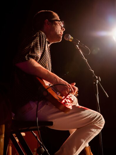 A musician sits on a stool and plays an instrument. He sings into the microphone and is illuminated on a stage. | © TVB Südsteiermark–Achromatic Photography