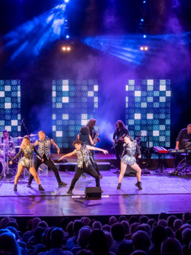 A live performance featuring multiple singers and dancers on stage. In the background are musicians and a colorful lighting design.