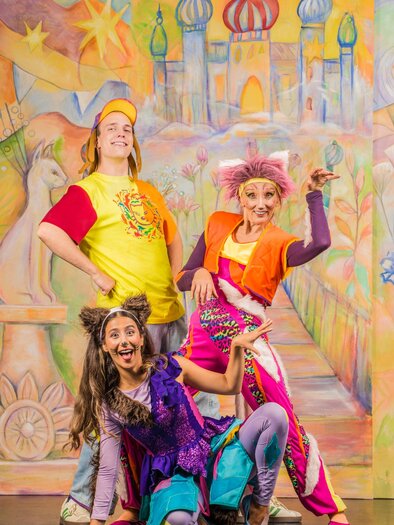 Drei fröhliche Schauspieler in bunten Kostümen posieren vor einer lebhaften Kulisse. Im Hintergrund sind Gebäude mit orientalischen Türmen und ein märchenhafter Weg zu sehen. | © Theater Heuschreck