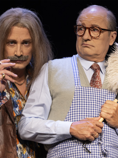 Two men on stage in humorous costumes. The man on the left has a mustached look and is smoking, while the other is holding a dust mop and looks surprised. | ©  Komödie Graz
