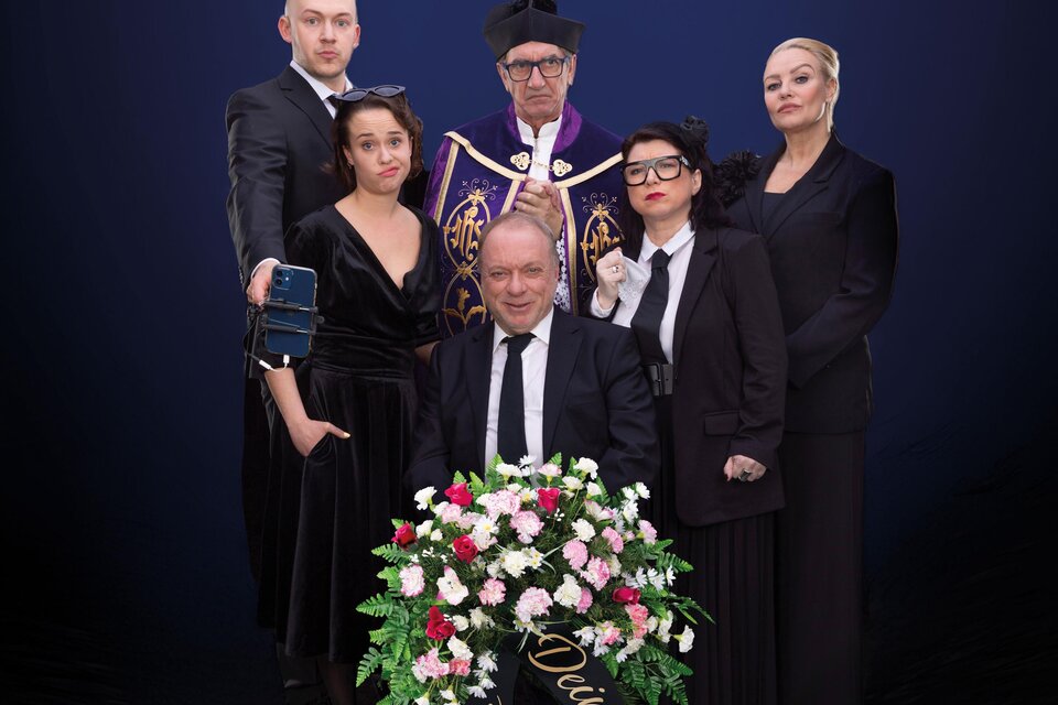 Eine Gruppe von sechs Personen in eleganter Kleidung steht zusammen vor einem dunklen Hintergrund. Die zentrale Figur hält einen Blumenkranz und es gibt eine feierliche Atmosphäre. | © Theater: Kalter weißer Mann
