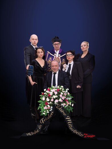 Eine Gruppe von sechs Personen in eleganter Kleidung steht zusammen vor einem dunklen Hintergrund. Die zentrale Figur hält einen Blumenkranz und es gibt eine feierliche Atmosphäre. | © Theater: Kalter weißer Mann