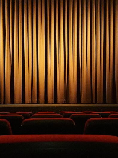 A dark theater with red seats and a closed curtain. The light illuminates the curtain in warm tones. | © Pixabay