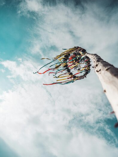 A tall mast with colorful ribbons reaches into the sky. In the background, bright clouds and a blue sky are visible. | © unsplash.com