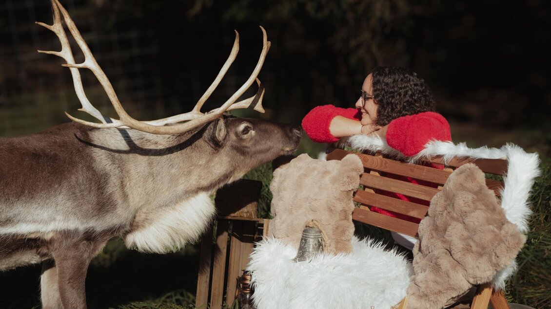 Eine Frau in einem roten Pullover sitzt auf einer Holzbänker, während ein rentierähnliches Tier neben ihr steht. Sie betrachten sich freundlich in einer natürlichen Umgebung. | © Einfachschön Fotografie