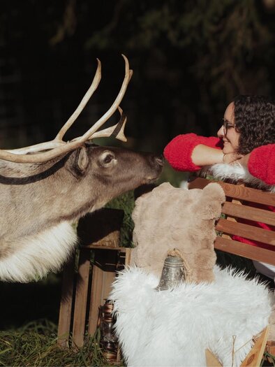 A woman in a red sweater sits on a wooden bench, while a reindeer-like animal stands next to her. They look at each other kindly in a natural setting. | © Einfachschön Fotografie