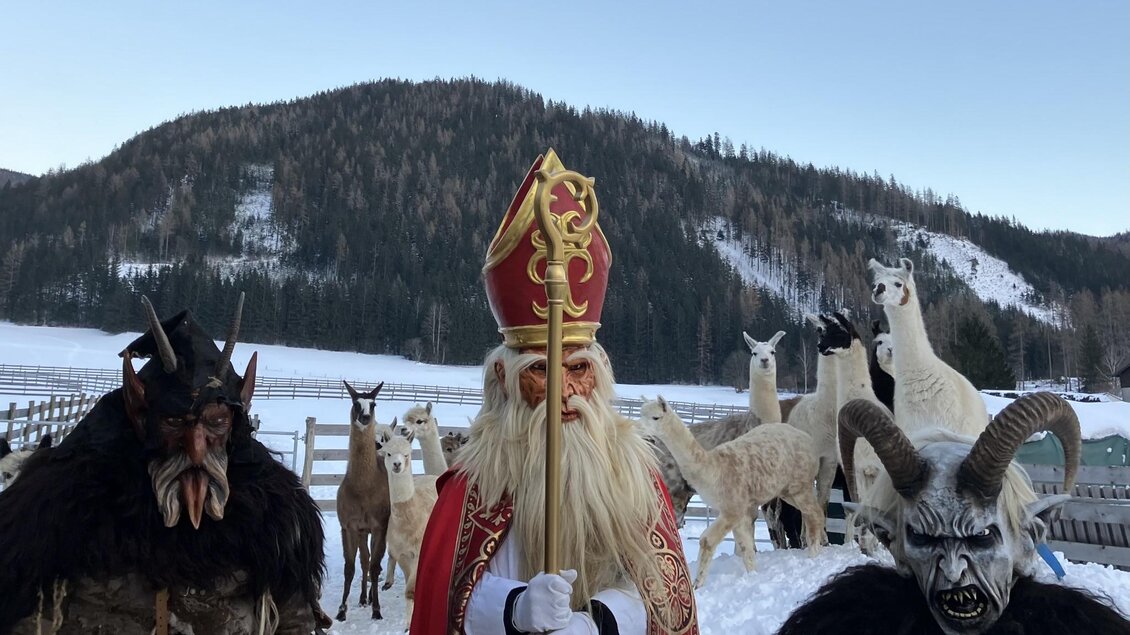 Eine winterliche Szene mit St. Nikolaus und zwei gruselig aussehenden Figuren in einem verschneiten Landschaftsraum. Im Hintergrund sind Lamas zu sehen, die in der Kälte stehen. | © Kaufmann