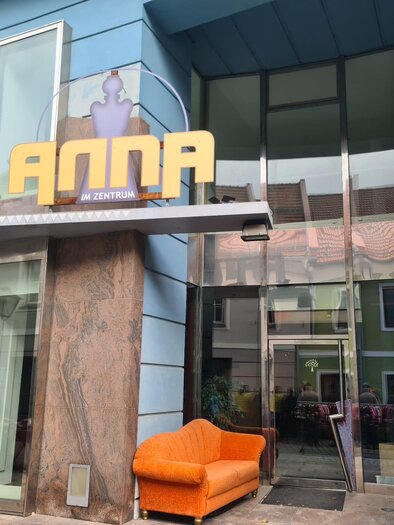 A modern building with the inscription "ANNA" and an inviting orange couch in front of the entrance. Next to the entrance, "CULTURAL ASSOCIATION MURAU" is written on the wall. | © Tv Murau