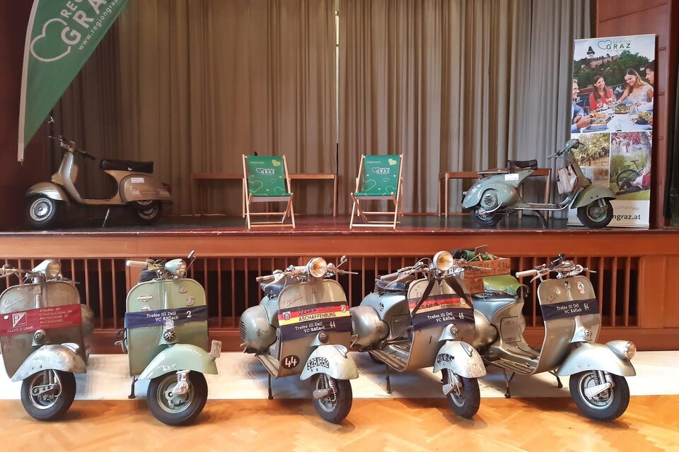 Eine Reihe von historischen Vespas steht auf einer Bühne. Im Hintergrund sind zwei grüne Stühle und ein Banner mit Informationen über die Region Graz zu sehen. | © Trofeo IV Del VC Köflach