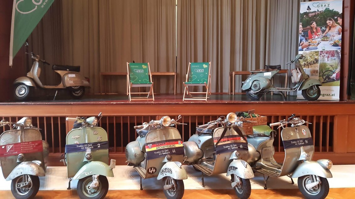 Eine Reihe von historischen Vespas steht auf einer Bühne. Im Hintergrund sind zwei grüne Stühle und ein Banner mit Informationen über die Region Graz zu sehen. | © Trofeo IV Del VC Köflach