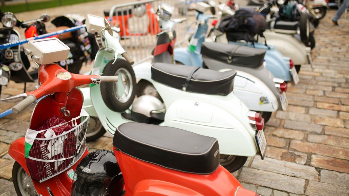 Eine Reihe von bunten Vespa-Rollern steht auf einem Pflasterweg. Im Hintergrund sind einige Motorräder zu sehen.