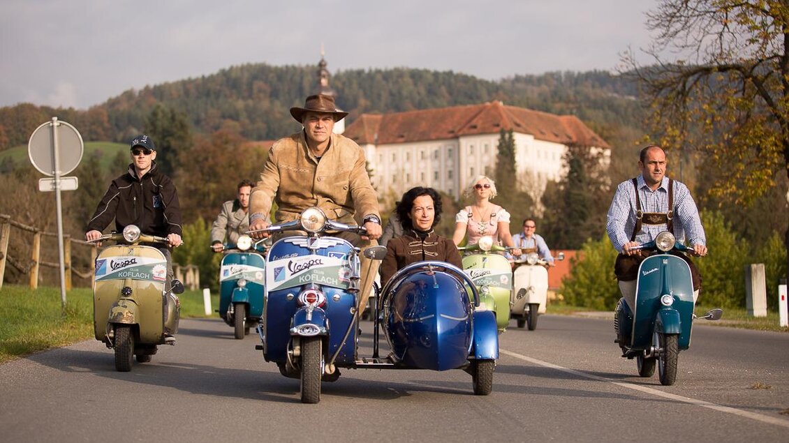 Eine Gruppe von Menschen fährt mit farbenfrohen Rollern auf einer Straße. Im Hintergrund sind Bäume und ein historisches Gebäude zu sehen. | © VideoFotograf.at