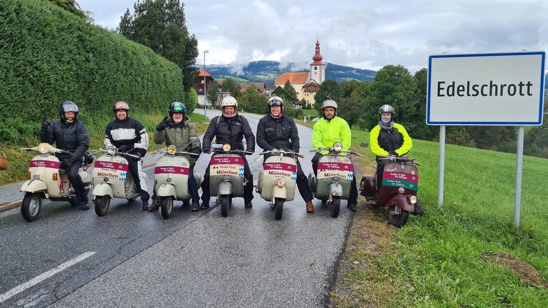 Eine Gruppe von acht Personen sitzt auf Rollern vor einem Ortsschild mit dem Namen "Edelschrott". Im Hintergrund sind Bäume und eine Kirche zu sehen. | © Trofeo IV Dell VC Köflach