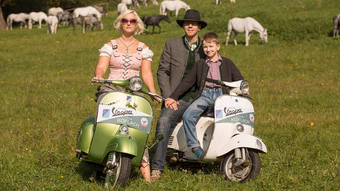 Ein Paar und ein Junge sitzen auf zwei farbigen Vespa-Rollern. Im Hintergrund sind grüne Wiesen und einige Pferde zu sehen. | © VideoFotograf.at