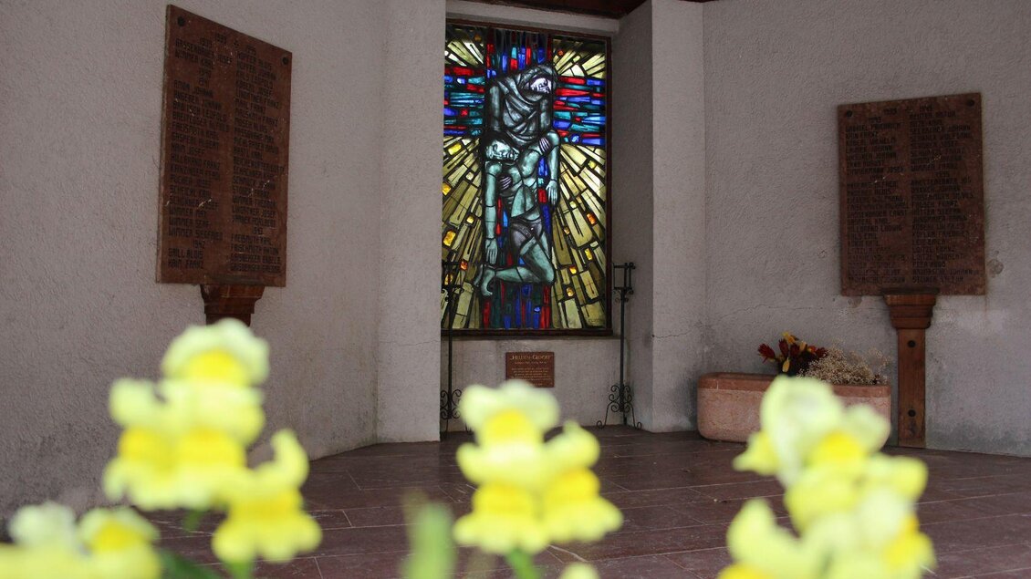 Ein kleiner Raum mit buntem Glasfenster und Blumen im Vordergrund. Es gibt zwei Wandtafeln aus Holz auf beiden Seiten. | © Viola Lechner