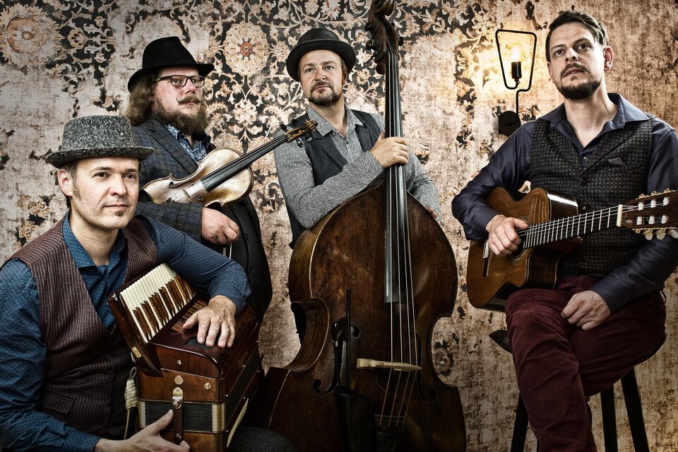 Eine Band mit vier Männern, die verschiedene Instrumente spielen. Sie stehen vor einer dekorativen Wand und lächeln in die Kamera. | © Tyrol Music Project