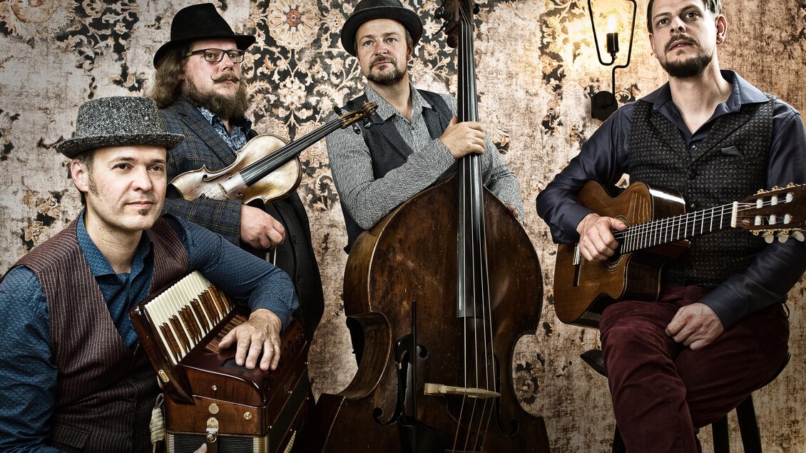Eine Band mit vier Männern, die verschiedene Instrumente spielen. Sie stehen vor einer dekorativen Wand und lächeln in die Kamera. | © Tyrol Music Project