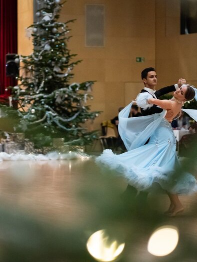 Ein Paar tanzt elegant in einem festlich geschmückten Raum. Im Hintergrund steht ein Weihnachtsbaum und Musiker spielen. | © Dance Unity
