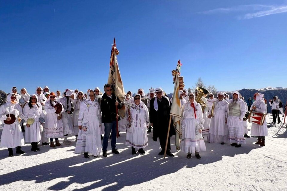Eine Gruppe von Menschen in traditionellen, weißen Kleidern steht auf Schnee unter einem strahlend blauen Himmel. Sie halten Fahnen und Musikinstrumente und scheinen an einer Feier oder einem Fest teilzunehmen. | © Martina Gaisberger