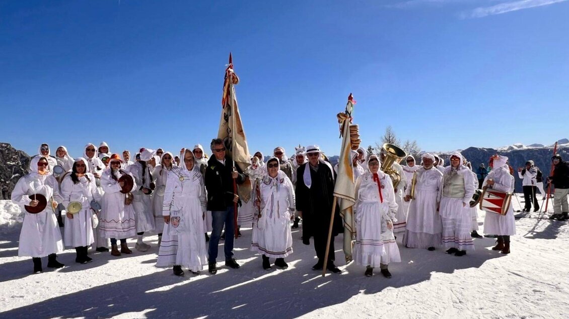Eine Gruppe von Menschen in traditionellen, weißen Kleidern steht auf Schnee unter einem strahlend blauen Himmel. Sie halten Fahnen und Musikinstrumente und scheinen an einer Feier oder einem Fest teilzunehmen. | © Martina Gaisberger