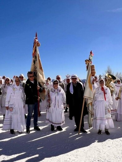 Eine Gruppe von Menschen in traditionellen, weißen Kleidern steht auf Schnee unter einem strahlend blauen Himmel. Sie halten Fahnen und Musikinstrumente und scheinen an einer Feier oder einem Fest teilzunehmen. | © Martina Gaisberger