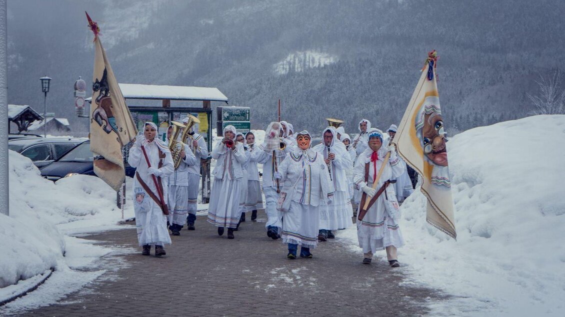 Eine Gruppe von Menschen in traditionellen weißen Kostümen marschiert durch eine verschneite Landschaft. Sie halten Fahnen und wirken festlich. | © Martina Gaisberger