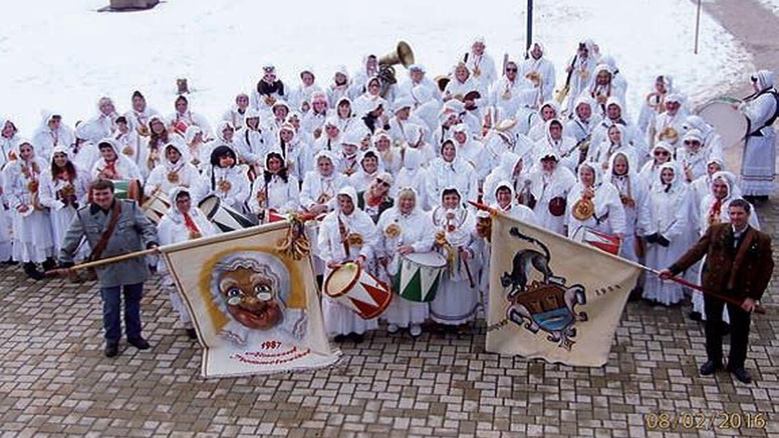 Eine große Gruppe von Menschen in weißen Kostümen versammelt sich draußen auf einem Platz. Einige halten Fahnen und Musikinstrumente, während Schnee am Boden liegt. | © Martina Gaisberger