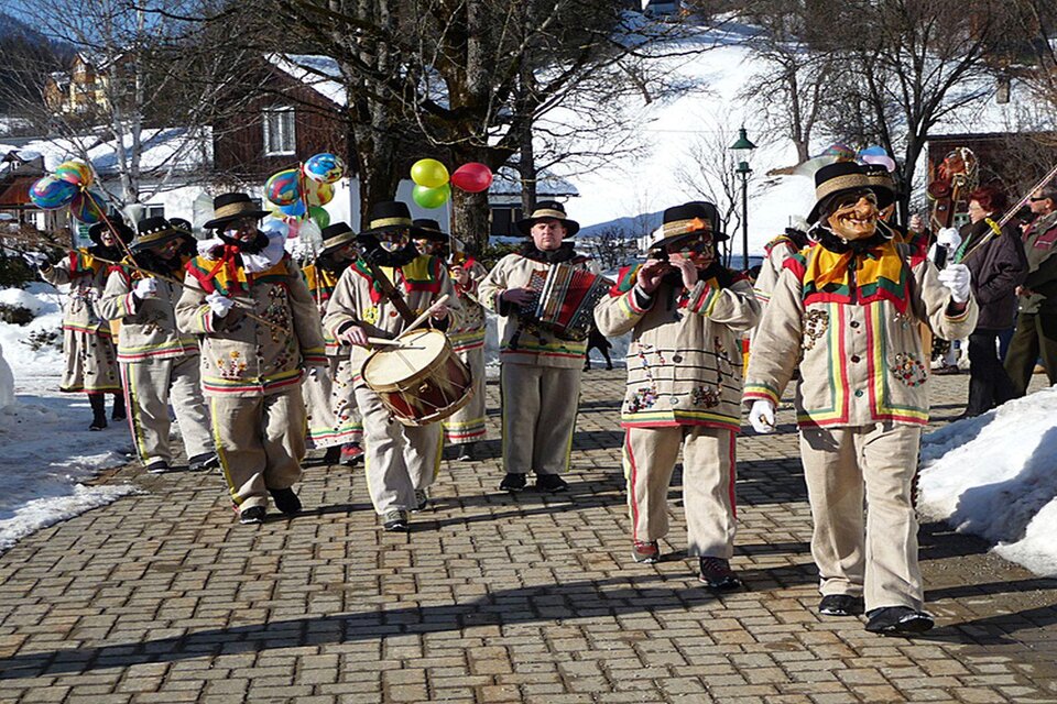 Eine fröhliche Gruppe von Menschen in traditionellen Kleidern, die durch eine schneebedeckte Straße marschiert. Sie spielen Instrumente und tragen bunte Girlanden und Luftballons. | © Hermann Schröttenhamer