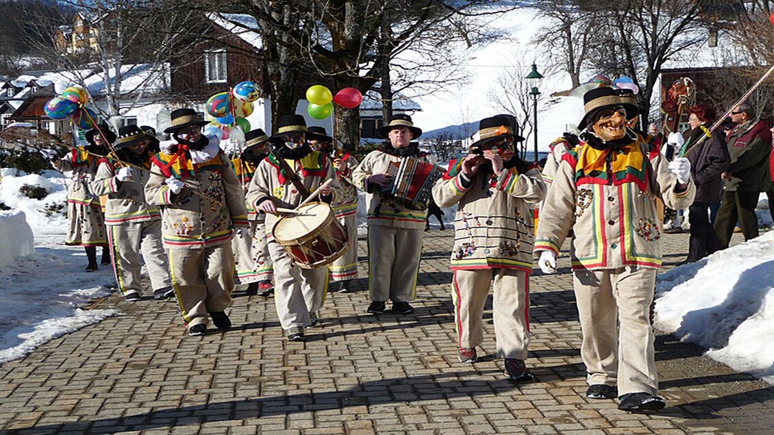 Eine fröhliche Gruppe von Menschen in traditionellen Kleidern, die durch eine schneebedeckte Straße marschiert. Sie spielen Instrumente und tragen bunte Girlanden und Luftballons. | © Hermann Schröttenhamer